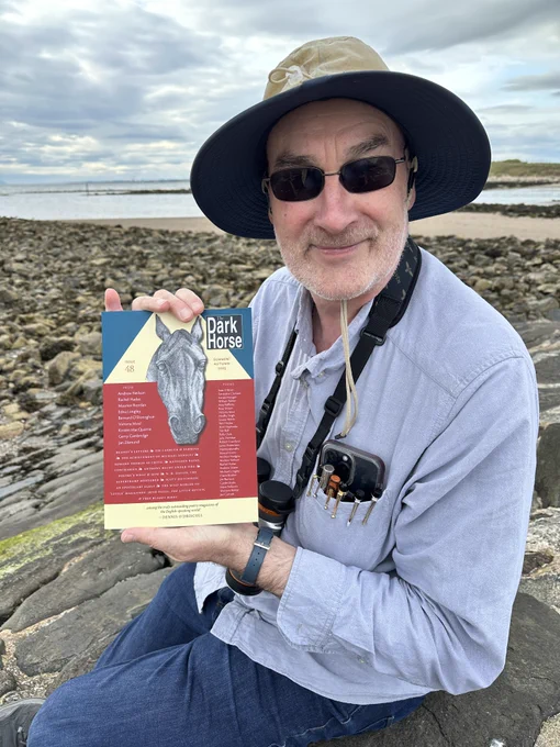Image of The Dark Horse's editor, Gerry Cambridge, in a pale grey shirt holding up a copy of the new issue, the whole framed against a sea background; he wears dark glasses and a wide brimmed sun-hat; he has fountain pens and a smartphone in his breast pocket; he has a pair of binoculars around his neck. He has a pleased smile on his face.