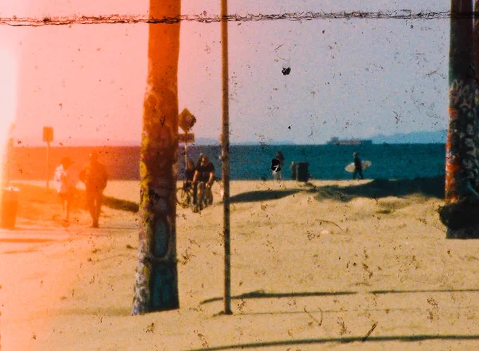 A still image from I Saw The Face Of God In The Jet Wash by Mark Jenkin depicting a beach scene with people walking, cycling and carrying a surfboard. Shot on grainy film.