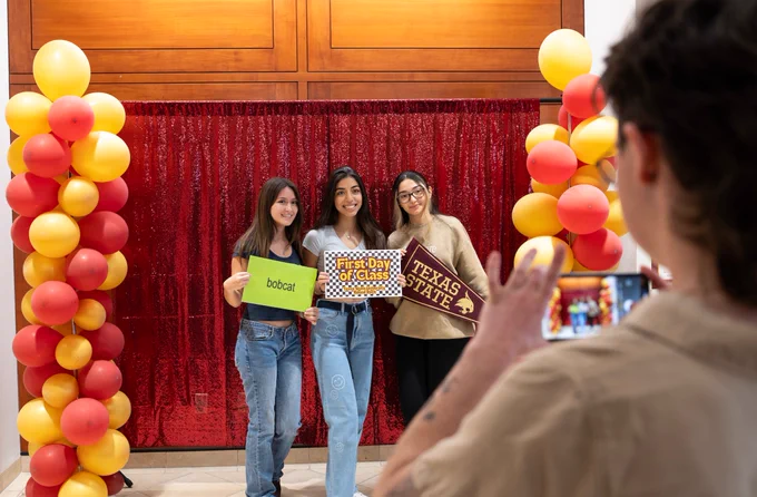 Three people stand in front of a red glittery backdrop, posing for a photo. They are holding signs that read "bobcat," "First Day of Class!," and "TEXAS STATE." The backdrop is flanked by columns of red and yellow balloons. Another person is taking their photo with a smartphone.