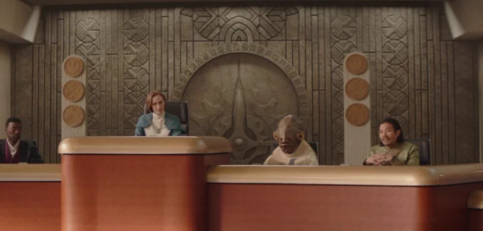 Mon Mothma and others sit behind a stand in a courtroom-like council chamber. The stone wall behind them is decorated with various markings and geometric patterns, including a great governmental seal which is surrounded by distinctive geometric Zeffo runes. From Ahsoka S1:E7.