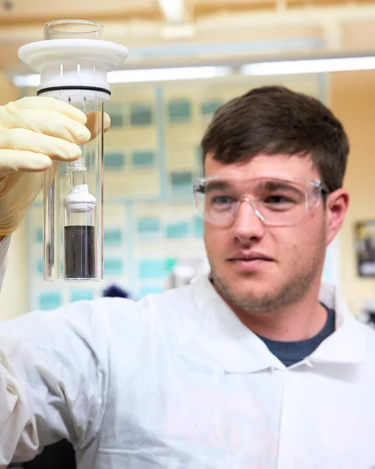 A laboratory technician wearing safety goggles and gloves is carefully examining a sample in a clear cylinder. The person is in a lab setting with equipment and cabinets in the background.