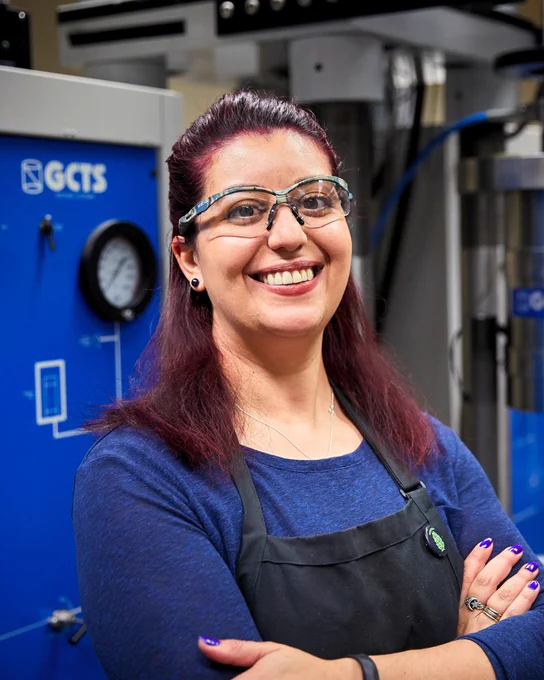 A person with red hair and glasses, smiling, wearing a dark apron stands in front of a blue GCTS machine in a laboratory setting.