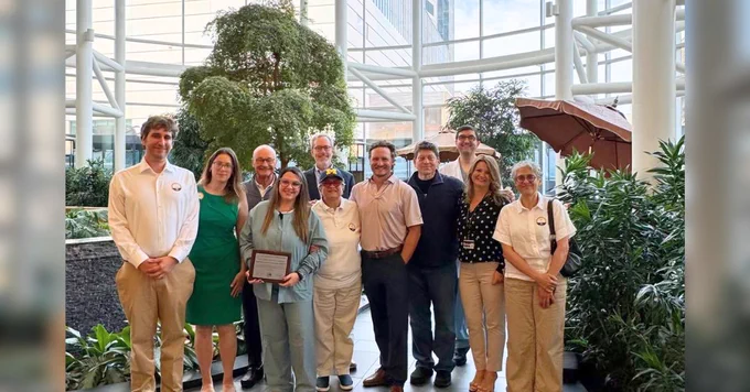 Group of eleven people posing with a certificate inside a brightly lit atrium with large glass panels and green plants.