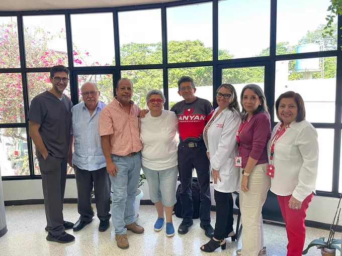 Group of seven medical staff and one patient smiling in a hospital lobby with large windows and flowering plants outside.