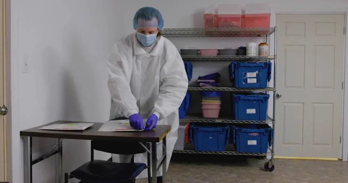 A person in a lab coat, gloves, and a face mask examines documents on a desk in a storage room with shelves filled with supplies.