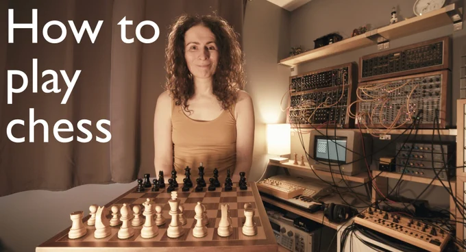 An overlaid title reads "How to play chess", over footage of me smiling over a chess board set up to play a game.  Much studio clobber is off to the side.  It's dark and cosy.