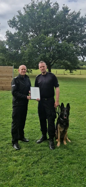 PD Bud, a black and tan German Shepherd is sitting down facing the camera.  Next to him on his right is PC Bendermacher dressed in police black uniform receiving the A4 sized framed certificate handed over by Ch Supt Dolan, also wearing police black uniform.  They are standing on grass with a tree behind them.