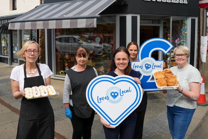 Deputy First Minister Kate Forbes standing outside of a shop. She is smiling and holding a sign which reads "Scotland Loves Local". She is joined by four women from Deas Bakery. Two of the women are holding trays of baked food. Another of women is holding a sign which reads "Scotland Loves Local".