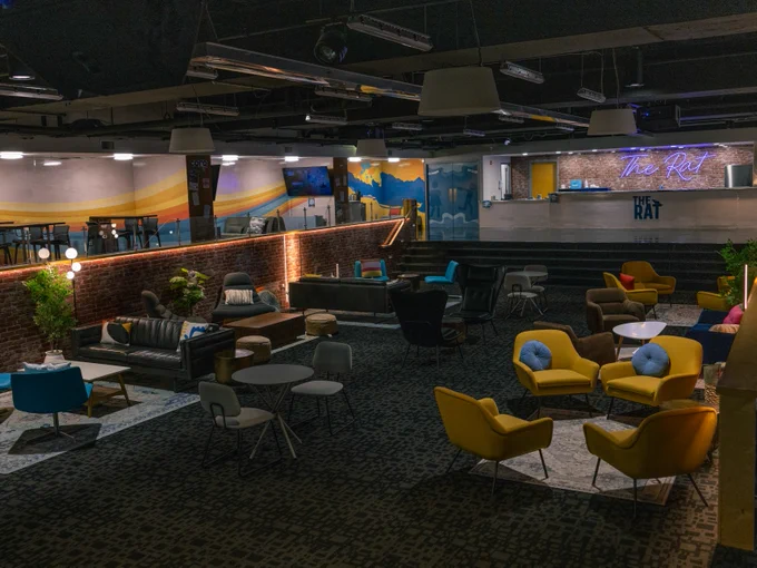 Interior view of The Rat, a modern lounge area with various seating arrangements including sofas and chairs, accent lighting, and decorative plants, set against a backdrop of wall murals and ambient lighting.