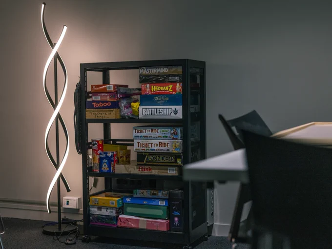 A dimly lit room with a bookshelf filled with board games including Mastermind, Taboo, Headbanz, Battleship, Ticket to Ride, and Risk, next to a modern, wavy floor lamp.