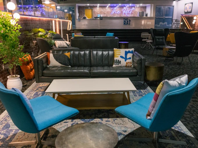 View of a lounge area at 'The Rat' featuring a large black sofa flanked by two blue chairs, a white coffee table, and decorative pillows. Dimly lit surroundings include more seating and tables in the background.