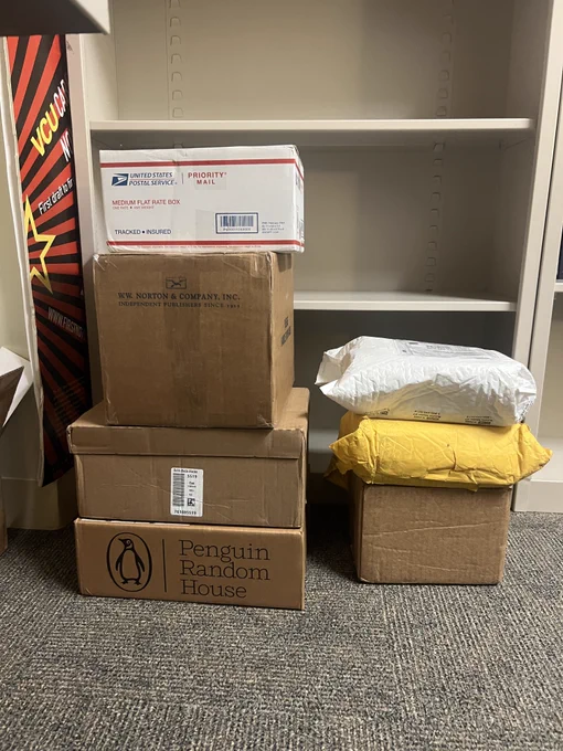 A pile of boxes sits on the ground in front of an empty bookshelf. 