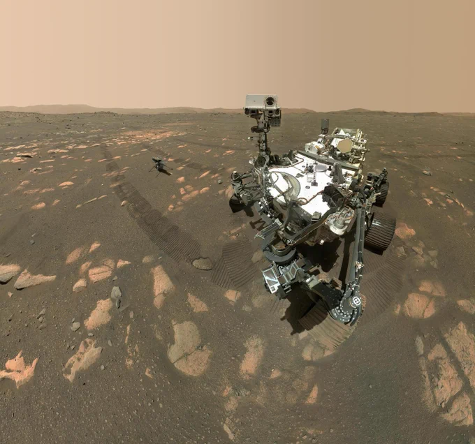 The SUV-sized Perseverance rover took this selfie on April 6, 2021, the 46th Martian day, or sol, of the mission, with its WATSON camera. The rover's tire tracks are visible proceeding into the distance. The small Ingenuity rotocopter is situated on the Martian surface behind and to the left of the rover inside Jezero Crater. Exposed orange-y rocks and dirt cover the surface. The sky is a greyish peach color.