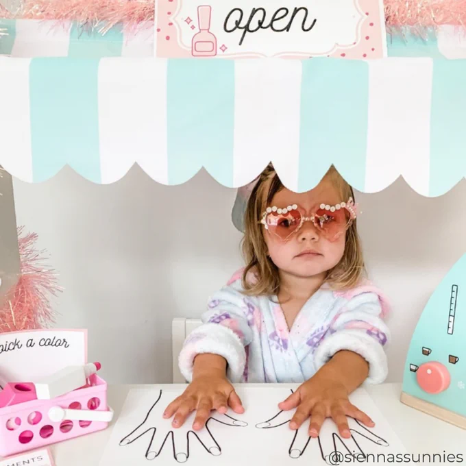 A child sits at a table with hand outlines on paper, wearing sunglasses with floral frames. Behind them is a playful setup resembling a manicure station with a sign that reads "OPEN" and various nail polish options displayed.