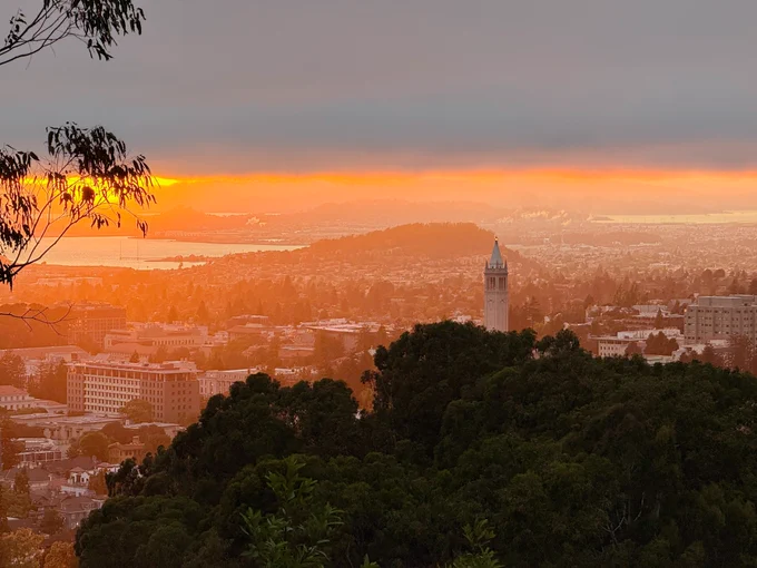 A sunset view from the Berkeley Hills overlooking the UC Berkeley campus and the San Francisco Bay. The Campanile tower peaks above hillside trees, and the setting sun casts a hazy orange glow over the scene under a layer of dark clouds.