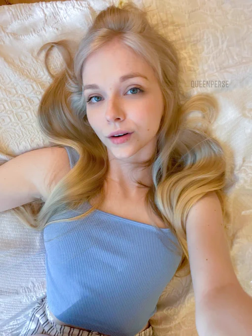 Queen Perse lying on a textured white blanket with her long hair spread around her face. She is wearing a light blue ribbed tank top and striped bottoms