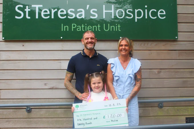 Young girl (Mollie) holding a cheque for one hundred and twenty pounds outside St Teresa's Hospice entrance. Her mum and dad are standing with her.