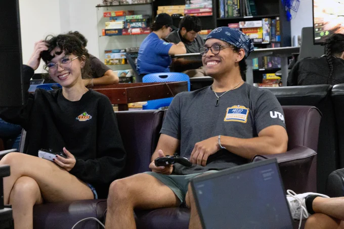 [ID: Pictured is a photo of two students playing a video game in The Basement of the UTA Central Library.]