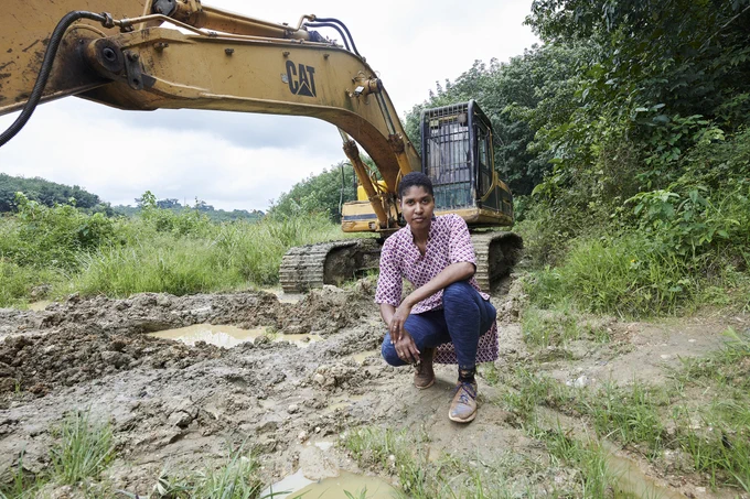 Professor Danielle Wood crouches in front of a large yellow excavator on muddy ground surrounded by lush greenery in Ghana. She wears a patterned shirt, blue pants, and boots, looking directly at the camera. The scene highlights deforestation and land disturbance caused by mining. Credit: Doug Segars