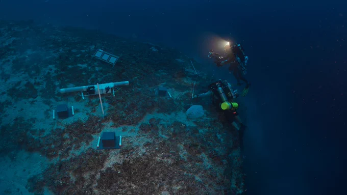 Two technical divers equipped with cameras and lights explore a mesophotic reef slope in dark waters in Fagatele Bay, American Samoa. Several scientific instruments are deployed on the seafloor.