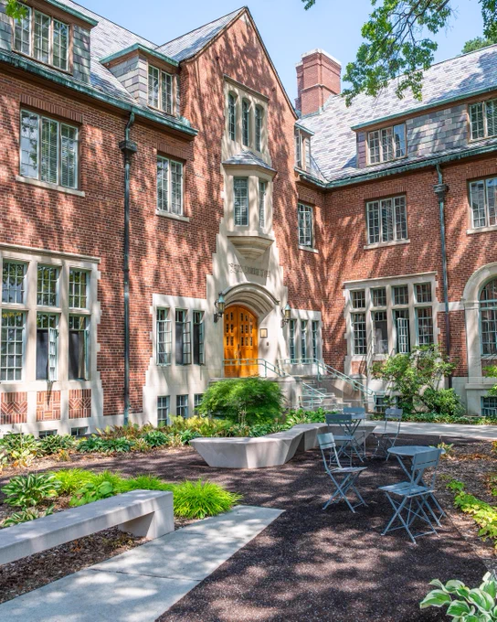 Exterior view of Yakeley, a brick structure with stone accents, featuring a courtyard with tables, chairs, plants, and a concrete bench. The building has multi-paned windows and a multi-tone slate roof.