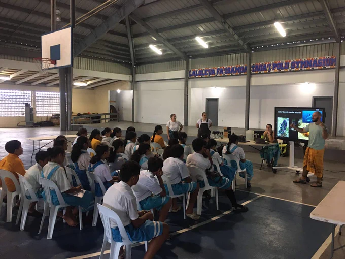 A group of students sits on chairs in a gymnasium, attentively listening to a marine science presentation delivered by educators at the front. 