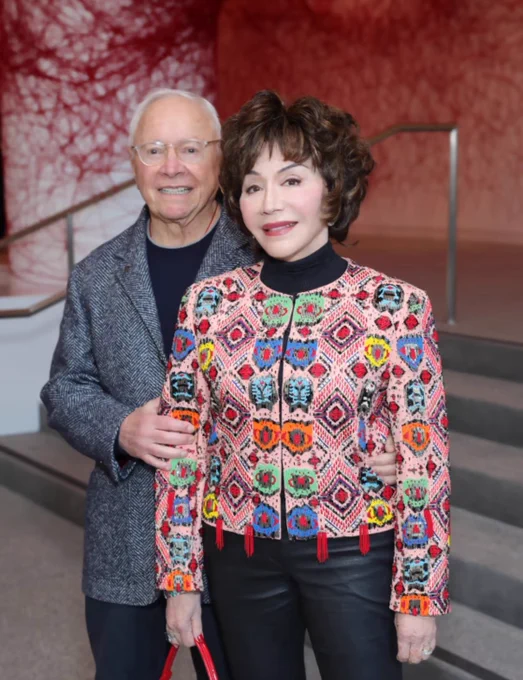 The billionaire couple posing. Stewart wears a grey jacket and Lynda wears a colorful jacket with black pants.