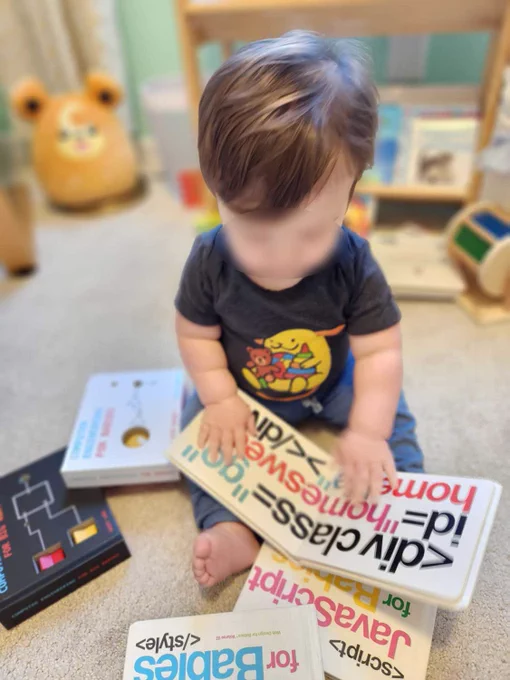 Really cute 10 month old baby with long brown hair and face blurred for privacy sits in the nursery wearing a gray Wapuu onesie. He is surrounded by books on coding for babies, and has HTML For Babies open in front of him.