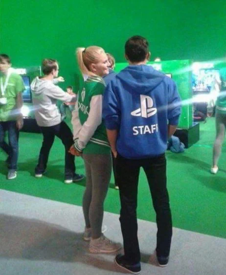 A photo shows an Xbox employee and a Playstation employee talking to each other at an event. The Xbox employee is wearing a green Xbox branded jacket and the Playstation employee is wearing a blue Playstation branded hoodie