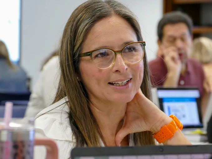 Person wearing glasses and an orange watch smiling during a meeting with a laptop and a beverage in front of them.
