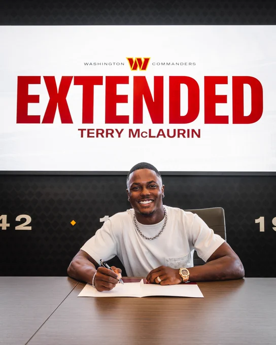Photo of Terry McLaurin singing a contract. He is smiling. Above him, on a screen, it says "EXTENDED" with "TERRY McLAURIN" underneath it. He is wearing a white shirt.