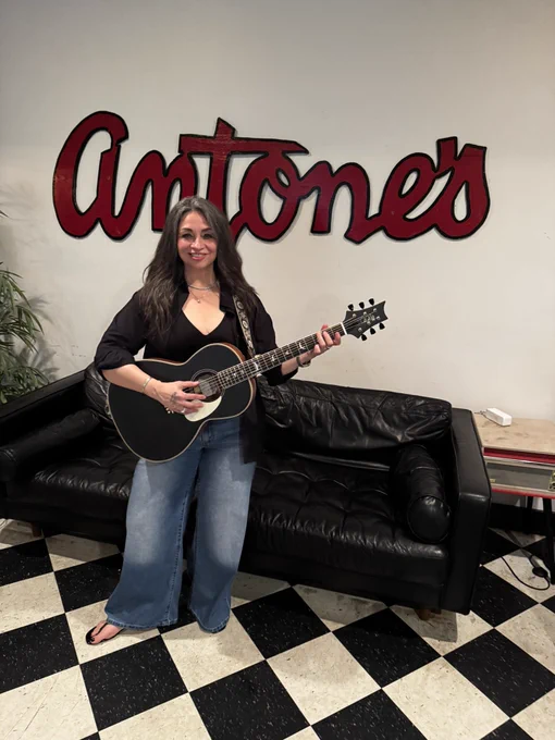 Diana standing with guitar, posing in front of black couch in Antone’s green room
