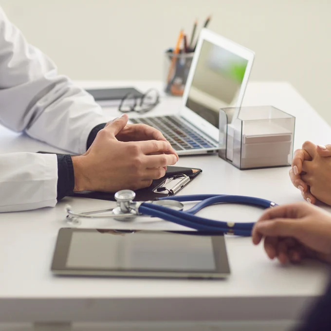 Doctor and patient sitting at a desk with a laptop, clipboard, and stethoscope during a medical consultation.