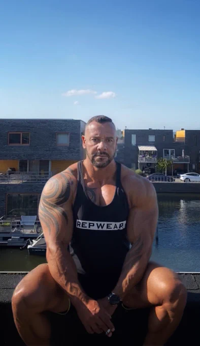 A powerful figure sits with arms resting on his legs, exuding strength and authority in a sleeveless black shirt.