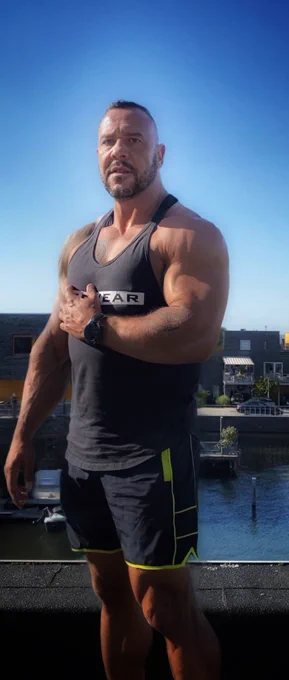 A heavily muscled man in a black tank top stands under a clear blue sky, his tattoos and build radiating dominance.