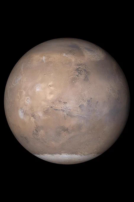 Color-enhanced Hubble Space Telescope image of Mars, showing its reddish-orange surface with dark patches, polar ice caps, and thin atmosphere against black space. Image courtesy NASA.