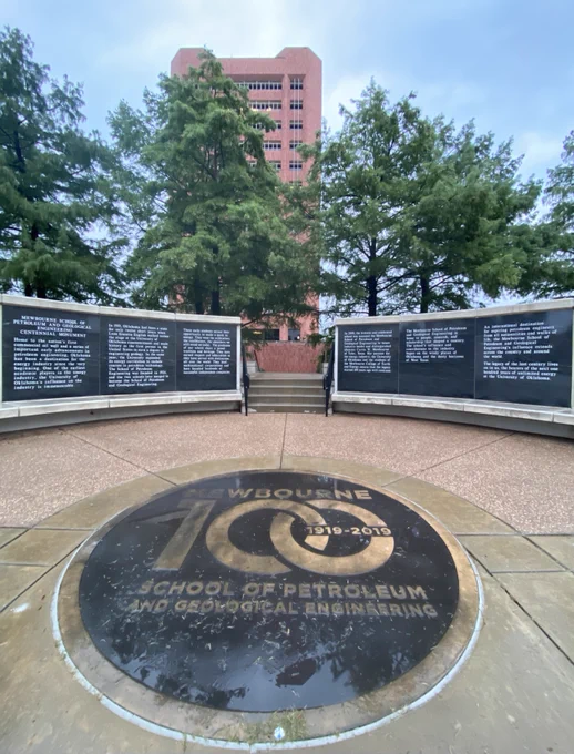 Photo of tall building (Sarkeys Energy Center on OU campus) and nearby trees.  In the foreground are several panels of dedicatory signage (including a gold and black seal on the ground), celebrating the 100th anniversary (1919-2019) of Mewbourne School of Petroleum and Geological Engineering. 