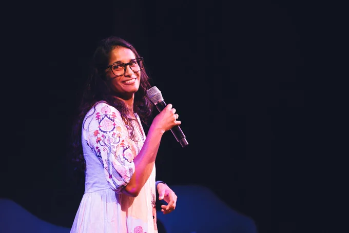 Tiffany holding a microphone on stage, dressed in a floral embroidered outfit, smiling during a presentation.