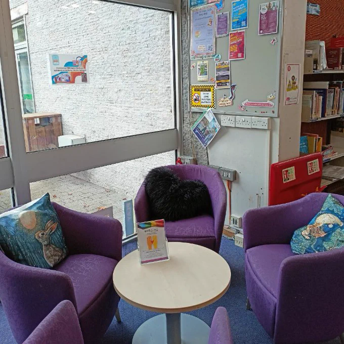 Photograph of the teen area with the noticeboard in the background and purple chairs with decorative cushions around wooden coffee table in the foreground. On the noticeboard leaflets promoting youth services as well as a poster for the Ewell Library Young Adult Book Club. On the window is a sign promoting eBooks and e-Audiobooks available from the Surrey Libraries Catalogue.