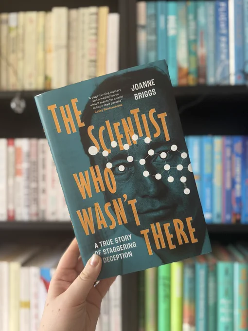 Hand holds a book titled "The Scientist Who Wasn't There" by Joanne Briggs. Background shows bookshelves with colorful books.