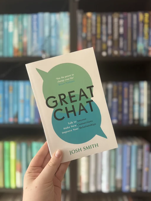 Hand holding the book "Great Chat" by Josh Smith, with a green speech bubble design. Bookshelves with colorful spines in the background.