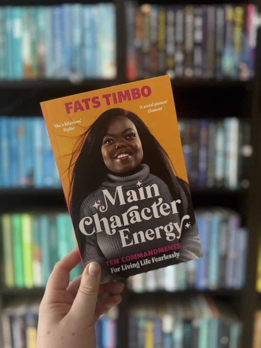 A hand holds a book titled "Main Character Energy" by Fats Timbo against a blurred bookshelf background. The cover is bright yellow-orange.