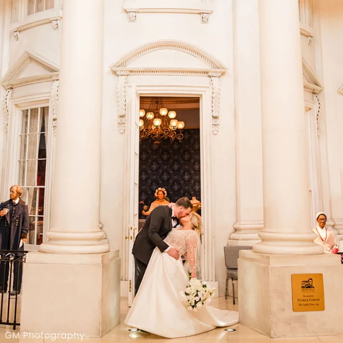 A bride and groom share a kiss in front of the White House replica inside the Abraham Lincoln Presidential Museum. The bride holds a bouquet of white flowers while wedding guests and life-sized figures look on from nearby.