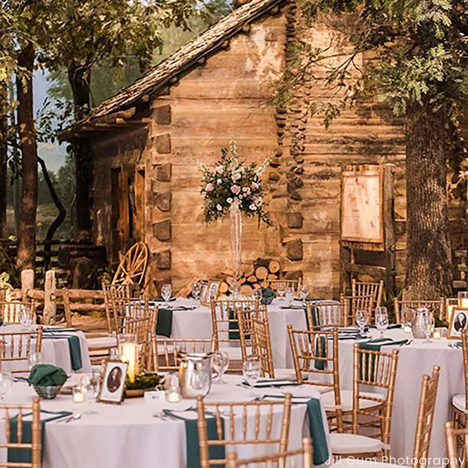 Round tables with white linens, green napkins, and gold chairs are set for a wedding reception in front of a rustic log cabin backdrop at the Abraham Lincoln Presidential Museum. Floral centerpieces and candlelight add elegance to the historic scene.