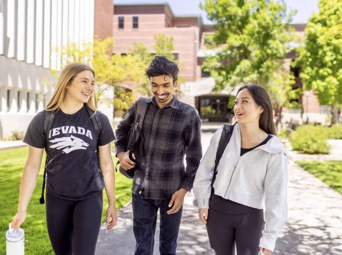 Three students, one wearing a Nevada t-shirt, walking and smiling on a sunny college campus.