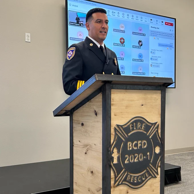 Joel Gallegos in a firefighter uniform standing at a podium with a badge logo that reads "BCFD 2020" in front of a screen displaying various icons.