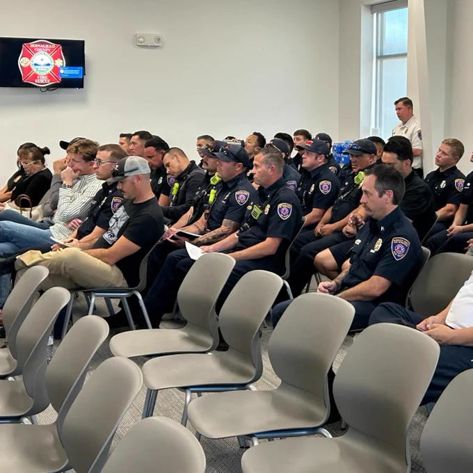 Group of uniformed firefighters and police officers seated during a training session in a classroom setting with a logo of Bernalillo County's fire department displayed on the screen.