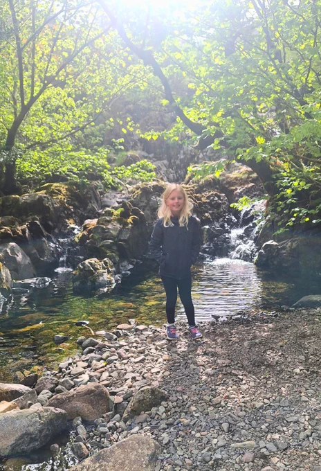 Emelia standing next to a river surrounded by trees. It is a very picturesque and tranquil scene.