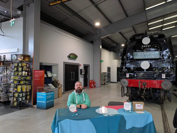A photo of Joe sitting behind a table with a Euan's Guide table cloth and marketing materials and Joe is wearing a spearmint Euan's Guide hoodie. There is a train in the background. 
