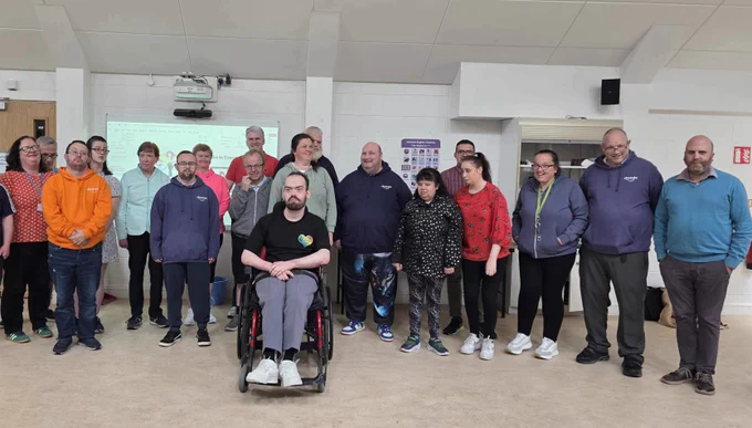 A group of disabled people, staff, and advocates are standing together at Sunbeam House Services for the relaunch of the Charter of Rights on Thursday, 28th August. One man, seated in a wheelchair, is at the front. NAS Aadvocates Ian McGahon and Ruth McCullagh are among the group. A presentation screen and a human rights poster are visible behind them.
